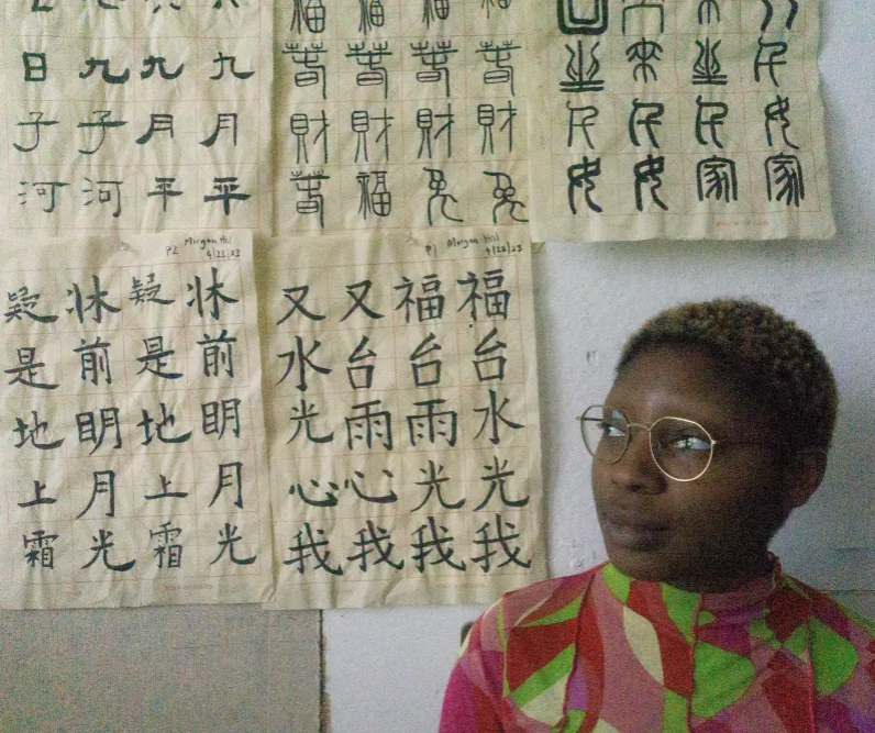 Morgan gazes to the left against the wall, with sheets of chinese calligraphy pinned to the wall behind her